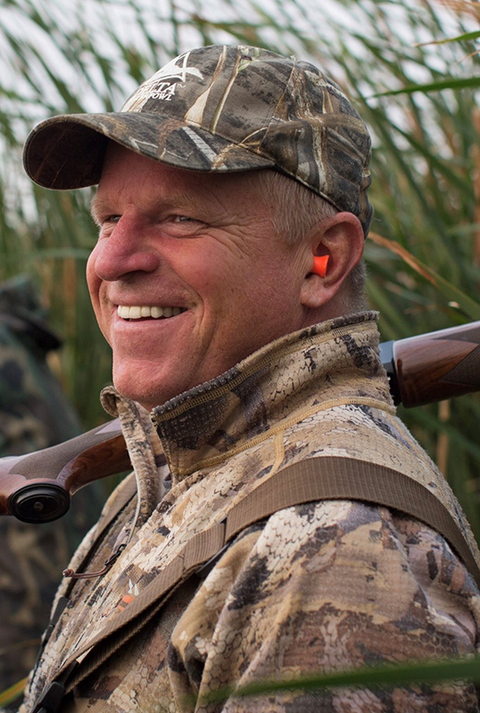 Dr. Scott Petrie is seen in his camo gear with hearing safety on among the long grass on a hunt.
