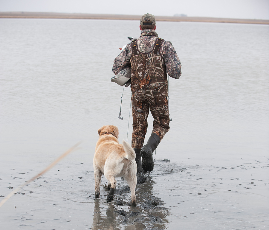 A solitary hunter and his dog walk through the grey mud of a slough on an overcast day.