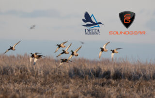 A flock of mallards flies over a brown field. The Delta Waterfowl and SoundGear logos can be seen.